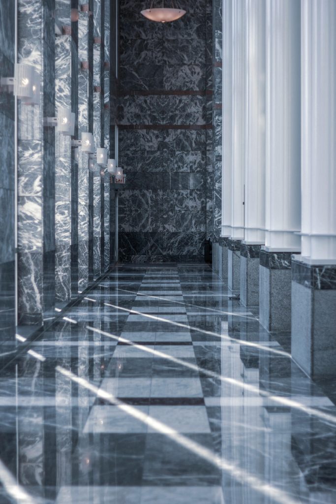 Modern marble hallway with tall columns and reflective surfaces illuminated by daylight.